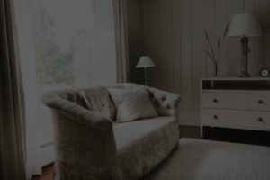 A cozy living room with a tufted beige loveseat, textured cushions, and Edson Hill charm—a white dresser with a lamp and vase, a small table lamp, and sheer curtains letting in natural light.