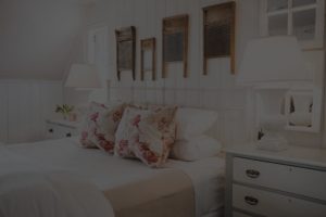 A cozy Edson Hill Guest Rooms bedroom with white walls, white bedding, floral pillows, two nightstands with lamps, and vintage washboards hanging as wall decor above the bed.