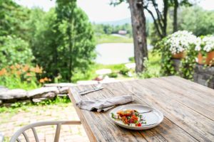 A plate of colorful food sits on a rustic wooden table outdoors, with a napkin and fork nearby. The background features green trees, flowers, and a pond under bright, natural light—perfect for a Vermont Vacation picnic.