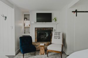 A cozy Edson Hill living room corner with a brick fireplace, flat-screen TV above it, blue velvet chair, cushioned armchair draped with a plaid blanket, small wooden stool, and built-in shelves displaying decor and books.