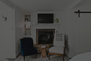 A cozy living room at Edson Hill with a brick fireplace, a TV mounted above, a dark blue armchair, a white armchair draped with a plaid blanket, a small wooden table, and shelves filled with books and décor.
