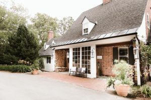 A charming brick house with a black roof, white trim, large front windows, and stacked firewood—perfect for a cozy Vermont vacation. Potted plants and greenery line the walkway, with a welcoming bench by the entrance.