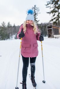 A woman with long blonde hair enjoys cross country skiing in the snow, wearing a pink coat, black leggings, gloves, and a blue winter hat with a pom-pom. Snow-covered trees and a wooden cabin are visible in the background.