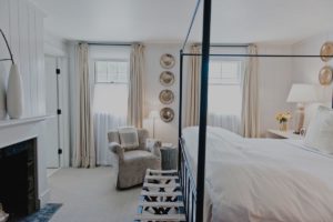 Bright, airy Edson Hill Guest Room with white walls and bedding, a large bed with a metal frame, a cozy armchair by the window, a fireplace, neutral curtains, and decorative plates on the wall.