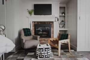 Cozy living room with an armchair, wooden chair, pouf, and fireplace inspired by Edson Hill. Both chairs have green plaid blankets. A TV is mounted above the fireplace, and built-in shelves hold books and small decor items.