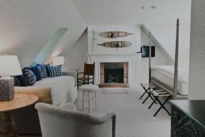 A cozy, modern attic bedroom in Edson Hill Guest Rooms features sloped ceilings, a brick fireplace, white linens, a sofa with blue pillows, an armchair, a small round table, and decorative snowshoes above the mantel.