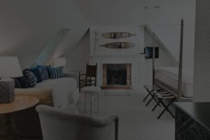 A cozy attic bedroom at Edson Hill Guest Rooms features sloped ceilings, a fireplace, a sofa, a bed, and rustic decor with snowshoes above the mantle. The softly lit space is decorated in calming neutral and blue tones.