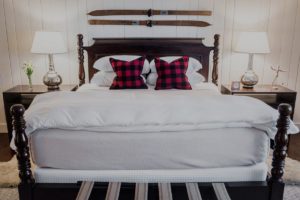 A neatly made bed with white bedding and two red and black plaid pillows, flanked by nightstands with lamps. Two vintage wooden skis are mounted on the white-paneled wall above the dark wood headboard, evoking cozy Edson Hill charm.