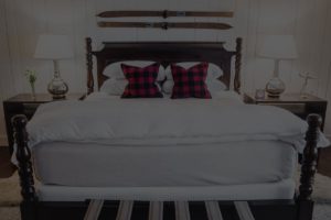 A neatly made bed with white bedding, a dark wooden frame, and two plaid throw pillows captures the cozy charm of Edson Hill. Two bedside tables with lamps flank the bed; vintage skis are mounted on the wall above the headboard.