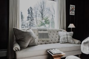 A cozy living room at Edson Hill features a cream-colored tufted sofa, large gray and plaid pillows, a striped throw blanket, a wooden coffee table with a tray, and a window framing snowy trees outside.