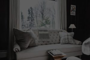 A bright living room at Edson Hill with a cream-colored tufted sofa, gray and patterned pillows, and a striped throw blanket. Large window behind the sofa reveals snowy trees outside. A small lamp and artwork are on the right.