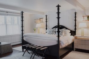 A cozy bedroom at Edson Hill features a black four-poster bed, crisp white bedding, patterned pillows, light wood nightstands with lamps, a bench at the foot of the bed, and a large window with sheer curtains letting in natural light.