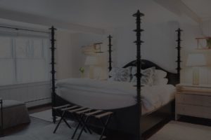 A cozy bedroom at Edson Hill features a four-poster bed with white bedding, two nightstands with lamps, a bench at the foot of the bed, and a window with sheer curtains letting in natural light.