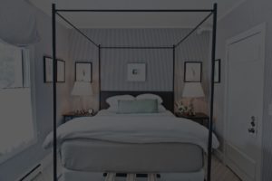 A neatly made canopy bed with white bedding and a light green pillow sits between two nightstands in a softly lit, gray walled bedroom. Framed artwork hangs above the bed, capturing the inviting charm of Edson Hill Guest Rooms.