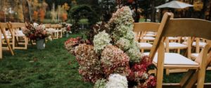 Outdoor weddings shine with rows of wooden chairs and large bouquets of white, pink, and green hydrangeas lining the aisle, while autumn trees create a stunning backdrop.