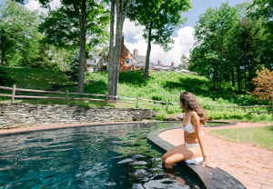 A woman in a white bikini sits on the edge of a round pool, dipping her feet in the water. She gazes toward Edson Hill - Stowe Vermont Inn, nestled among trees and greenery on a bright, sunny day.