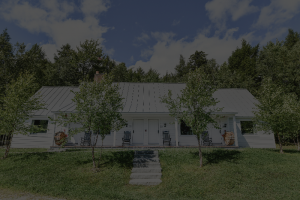 A charming white, single-story house with a metal roof and rocking chairs on the front porch offers a classic Vermont Vacation vibe, surrounded by potted plants, green trees, and grass under a partly cloudy sky.
