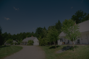 A row of small white cottages with porches sits beside a gravel path, surrounded by green grass and trees under a clear blue sky—perfect for a peaceful Vermont vacation.