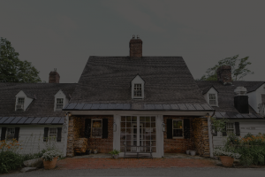 A large, traditional house with a steep, dark roof, central brick chimney, white walls, and brick entryway—perfect for a relaxing Vermont vacation. Potted plants and stacked firewood accent the glass front door, with trees in the background.