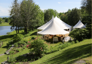 White event tents are set up on a grassy hillside at Edson Hill - Stowe Vermont Inn, surrounded by lush green trees and bushes, with a pond and blue sky in the background. Tables and chairs under the tents suggest a festive gathering or celebration.