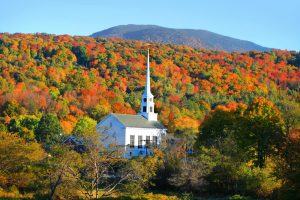 A white church with a tall steeple stands among vibrant Vermont Fall Foliage, surrounded by colorful autumn trees and a forested mountain under a clear blue sky.