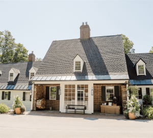 A charming brick and white cottage with a steep, dark shingled roof, a central chimney, large front windows perfect for watching for packages, and a shaded entrance adorned with potted plants and outdoor furniture.