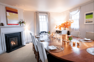 A cozy dining room with a long wooden table set for a meal evokes the elegance of fine dining in Stowe VT. Eight chairs, candles, glassware, plates, and orange autumn leaves as a centerpiece glow by the fireplace and modern art in this softly lit room.