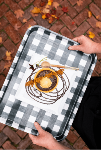A person holds a checkered tray with a plated dessert, featuring a round pastry topped with ice cream, chocolate drizzle, and garnishes. Perfect for enjoying Fine Dining in Stowe VT, autumn leaves and brick pavement are visible below.