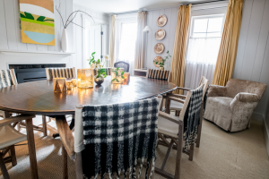 A cozy dining room reminiscent of fine dining in Stowe VT features a round wooden table with candles and plants, chairs draped in black and white checkered throws, light yellow curtains at the windows, and a beige armchair in the corner.