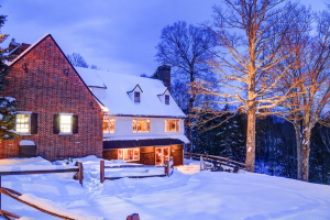 A cozy brick house with glowing windows sits surrounded by deep snow and bare trees at dusk, lights illuminating the wintry scene near a wooden fence—evoking the charm of Edson Hill - Stowe Vermont Inn.