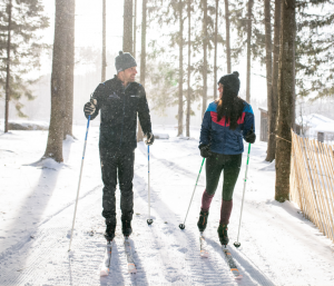 Two people dressed in winter clothing are cross-country skiing on a snowy trail through a forest, smiling at each other as sunlight filters through the trees, enjoying one of the resort’s winter adventure packages.