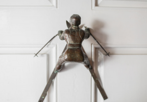 A metal door knocker shaped like a skier in mid-action, mounted on a white wooden door at Edson Hill - Stowe Vermont Inn. The skier’s body faces outward, with ski poles and legs spread apart, giving a dynamic appearance.