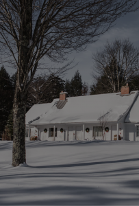 A house with a snow-covered roof sits in a snowy yard at Edson Hill - Stowe Vermont Inn, surrounded by bare trees. Wreaths hang on the windows, and shadows from trees stretch across the snow in the foreground.