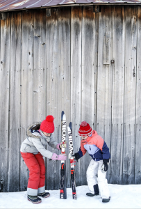 Two children wearing winter clothes and red hats stand in snow, holding upright skis together in front of a weathered wooden wall at Edson Hill - Stowe Vermont Inn.