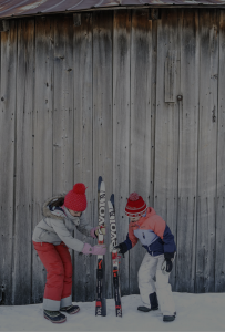 Two children in winter clothes and red hats stand in snow, arranging skis together in front of a weathered wooden wall at Edson Hill - Stowe Vermont Inn.