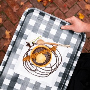 A hand holds a checkered tray with a dessert: a tart topped with ice cream, chocolate drizzle, and garnishes, on a square plate. Autumn leaves and a brick path set the scene for fine dining in Stowe, VT.