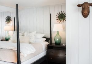 A cozy Edson Hill - Stowe Vermont Inn bedroom with a four-poster bed, crisp white bedding, green glass lamps on dark nightstands, sun-shaped mirrors on white-paneled walls, and a decorative animal head mounted above.
