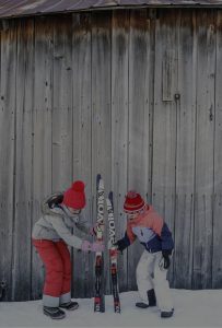 Two children in winter clothing and red hats stand in snow by a wooden wall, holding and arranging two pairs of skis upright between them, capturing the cozy charm of Edson Hill - Stowe Vermont Inn.