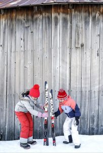 Two children in winter clothes and red hats stand in the snow, propping up skis together in front of a weathered wooden wall at Edson Hill - Stowe Vermont Inn.