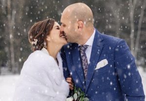 Bride and groom dressed warmly, smiling and touching noses in falling snow at Edson Hill - Stowe Vermont Inn, with a wintry backdrop of trees. The groom wears a blue suit while the bride, in a white shawl, holds a bouquet.