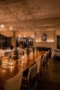A warmly lit restaurant with cozy decor, including candles, small pine trees, and fairy lights on a long wooden table. Two people chat in the background, creating a welcoming, intimate atmosphere perfect for casual fine dining.