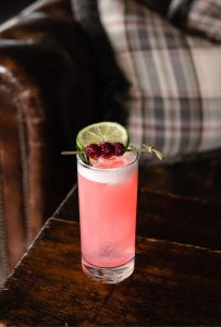 A tall glass of pink cocktail with ice, featured on the menu, is garnished with a lime wheel and red berries on a skewer. It sits on a wooden table beside a brown leather chair with a plaid-patterned cushion in the background.