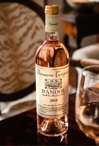 A bottle of Domaine Tempier Bandol 2019 rosé wine sits on a table, with a glass of wine and elegant, upholstered chairs in the background—an inviting scene perfect for any menu. The bottle’s label is clearly visible and detailed.