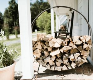 A metal firewood holder filled with chopped logs sits on a brick porch next to a potted plant and some packages outside a white house, with rocking chairs and greenery in the background.