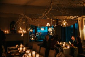 A warmly lit dining room with candles on the table and glass ornaments hanging from white branches above, creating a cozy, intimate atmosphere—perfect for a Thanksgiving Dinner in Stowe Vermont, with loved ones gathered in the background.