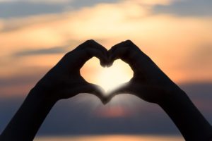 Two hands form a heart shape against a bright sunset sky, with sunlight shining through the center of the heart.