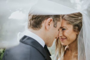 A bride and groom stand close together, touching foreheads and smiling gently, with a white veil softly draped around them, creating an intimate and romantic moment.