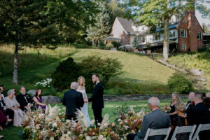 A bride and groom stand holding hands during an outdoor wedding ceremony, surrounded by seated guests and floral arrangements, with a large house and greenery in the background.