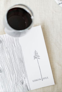 A glass of red wine sits next to a white card on a beige tablecloth. The card, evoking the warmth of a Thanksgiving Dinner in Stowe Vermont, features a simple tree illustration beside wood grain lines and “EDSON HILL” at the bottom.