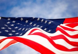 Close-up of the United States flag waving in the wind, showing white stars on a blue field and red and white stripes, with a background of a blue sky and light clouds.